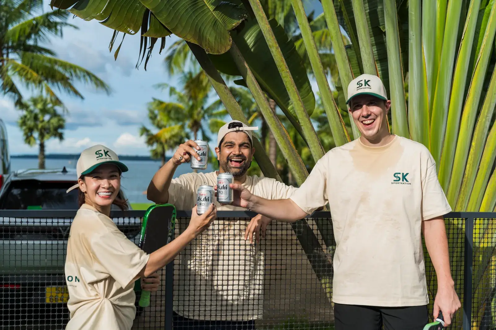 Sportkation participants raising drinks at the pickleball net beneath tropical trees